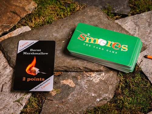 Education Outdoors S'mores the Card Game with green card stack and 2 point burnt marshmallow card against stone path and grass.