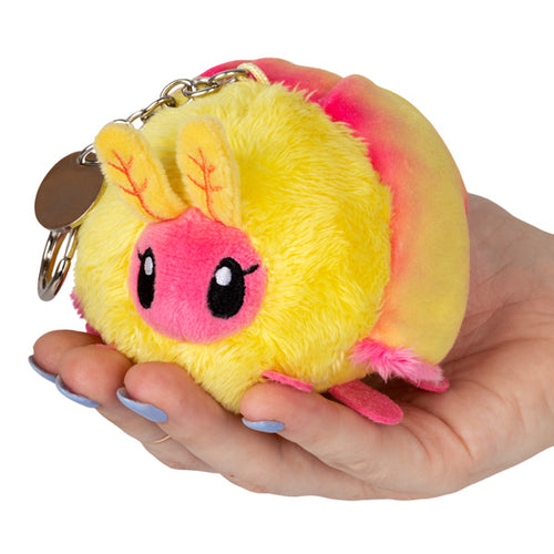 Squishables neon yellow and pink fluffy round body with eyelashes Rosy Maple Moth Keychain with silver chain with latch being held by a hand with blue nail polish aganist a white background.
